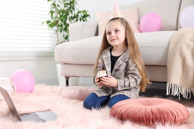 Photo of Birthday celebration. Little girl with cupcake having video call via laptop at home