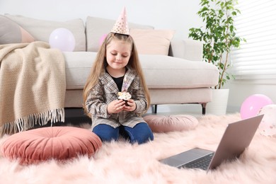 Photo of Birthday celebration. Little girl with cupcake having video call via laptop at home