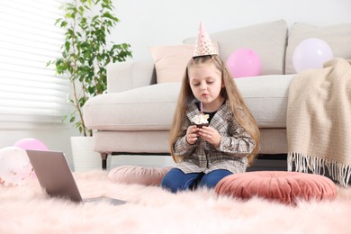 Photo of Birthday celebration. Little girl blowing candle on cupcake while having video call via laptop at home