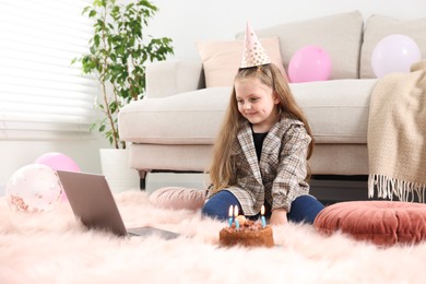 Photo of Birthday celebration. Little girl with cake having video call via laptop at home