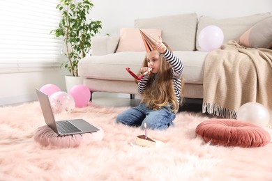 Photo of Birthday celebration. Little girl with blower and piece of cake having video call via laptop at home
