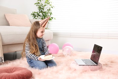 Photo of Birthday celebration. Little girl with piece of cake having video call via laptop at home