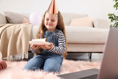 Photo of Birthday celebration. Little girl with piece of cake having video call via laptop at home