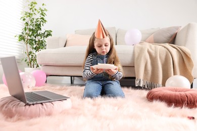 Photo of Birthday celebration. Little girl blowing candle on piece of cake while having video call via laptop at home