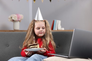 Photo of Birthday celebration. Little girl with piece of cake having video call via laptop on sofa at home