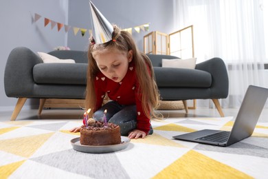Photo of Birthday celebration. Little girl blowing candles on cake while having video call via laptop at home