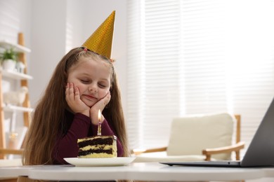 Photo of Birthday celebration. Little girl with piece of cake having video call via laptop at table indoors