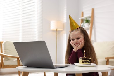 Photo of Birthday celebration. Little girl with piece of cake having video call via laptop at table indoors