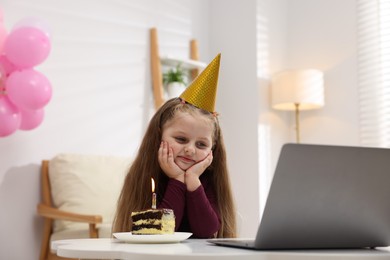 Photo of Birthday celebration. Little girl with piece of cake having video call via laptop at table indoors