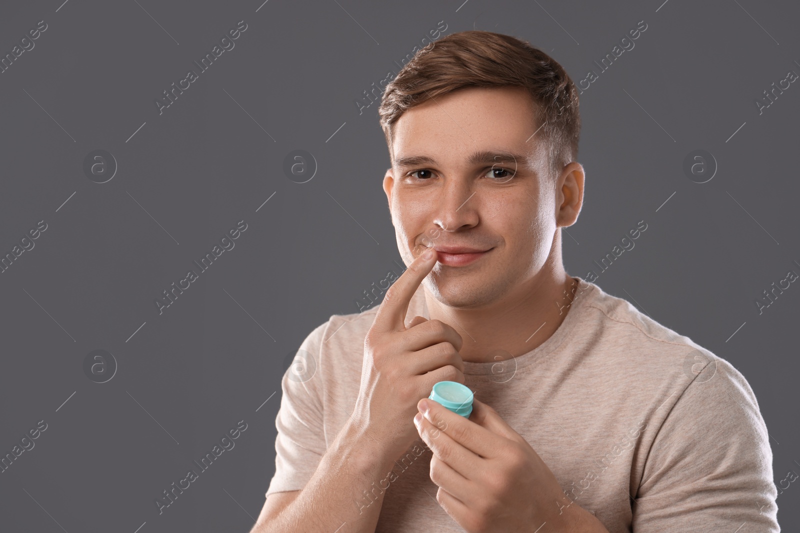 Young man applying lip balm onto his lips on grey background, space for text Photo of Young man applying lip balm onto his lips on grey background, space for text