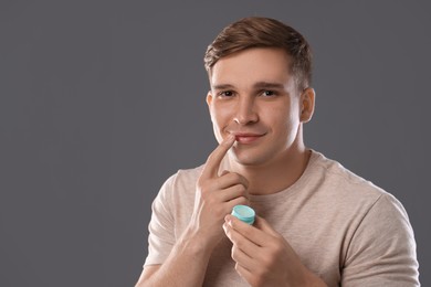 Photo of Young man applying lip balm onto his lips on grey background, space for text
