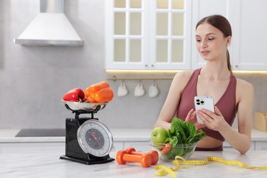 Photo of Woman using smartphone near white marble table with kitchen scale, healthy food, dumbbells and measuring tape indoors