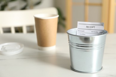 Photo of Receipt in metal bucket and paper cup of drink on white marble table in cafe, closeup