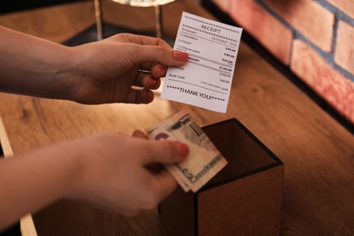 Photo of Woman with receipt putting tips into box at wooden table indoors, closeup