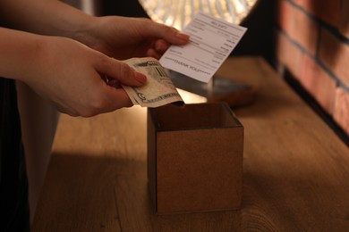 Photo of Woman with receipt putting tips into box at wooden table indoors, closeup