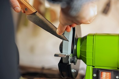 Photo of Man sharpening knife with bench grinder indoors, closeup