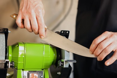 Photo of Man sharpening knife with bench grinder indoors, closeup
