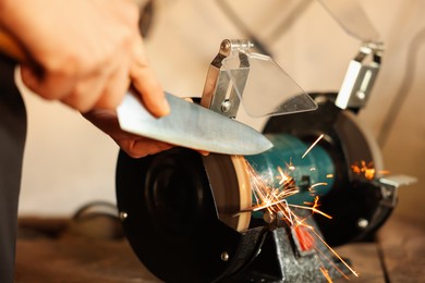 Photo of Man sharpening knife with bench grinder indoors, closeup