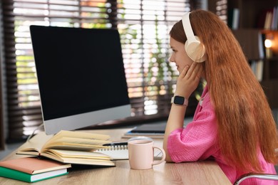 Photo of Teenage student with headphones using computer while studying at table indoors