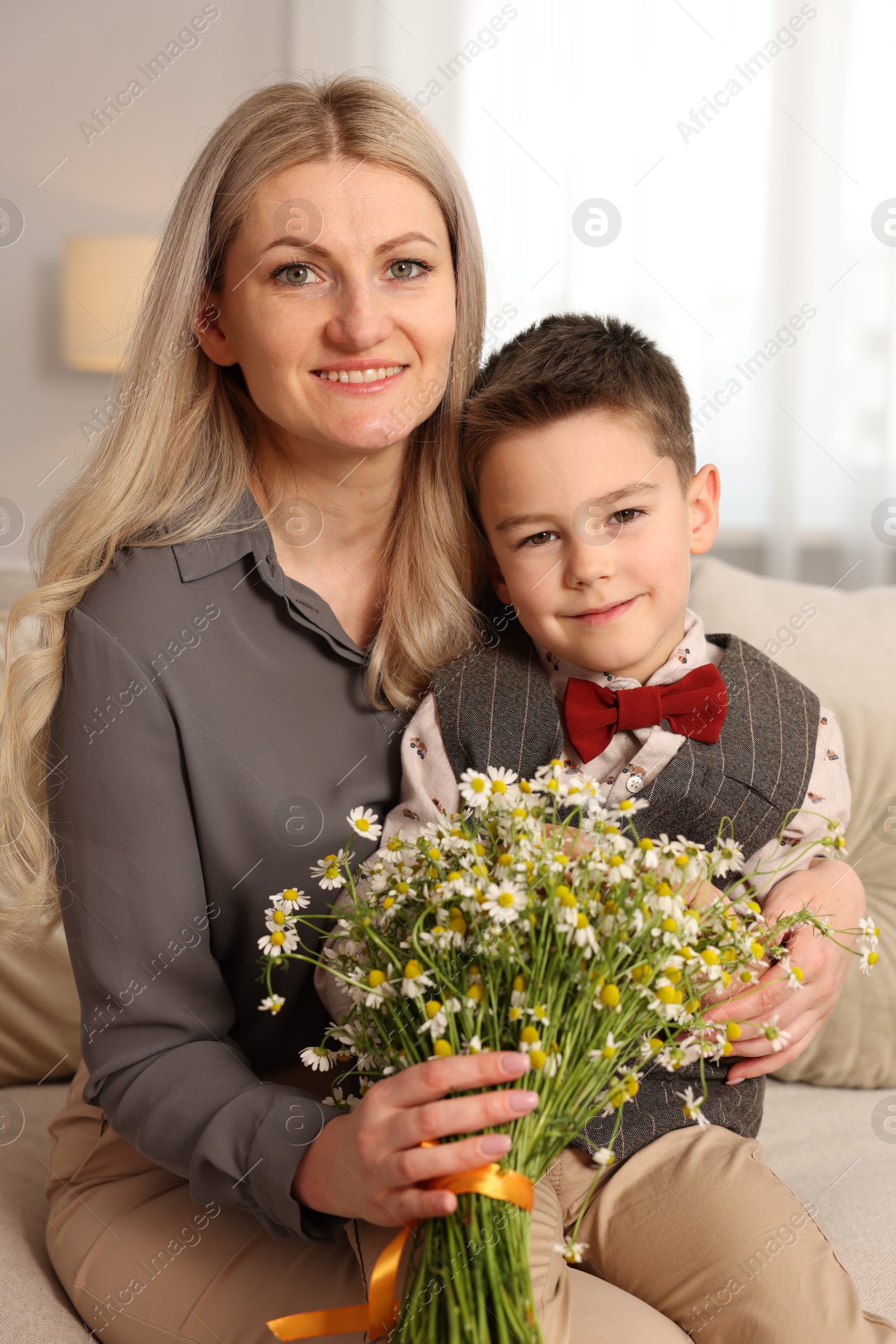 Little boy and his smiling mother with bouquet of beautiful chamomile flowers on sofa at home Photo of Little boy and his smiling mother with bouquet of beautiful chamomile flowers on sofa at home