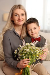 Photo of Little boy and his smiling mother with bouquet of beautiful chamomile flowers on sofa at home