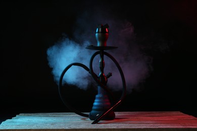 Photo of Hookah on wooden table with smoke in color lights against black background