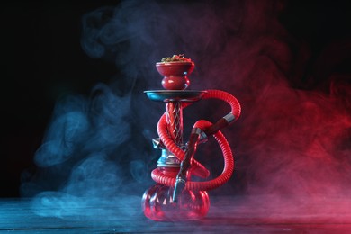Photo of Hookah on wooden table with smoke in color lights against black background