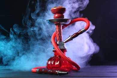 Photo of Hookah on wooden table with smoke in color lights against black background