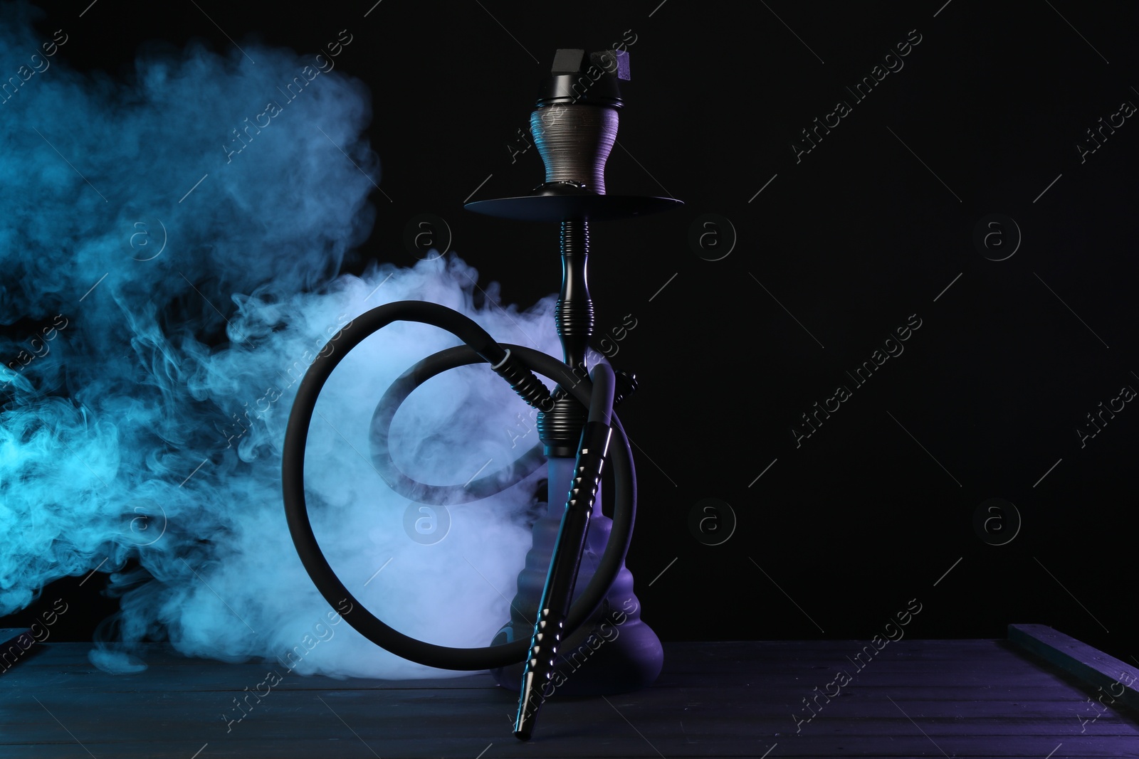 Photo of Hookah on wooden table with smoke in color lights against black background