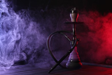 Photo of Hookah on wooden table with smoke in color lights against black background