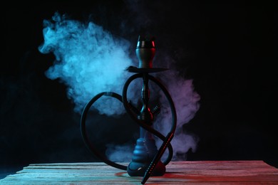 Photo of Hookah on wooden table with smoke in color lights against black background