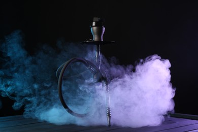Photo of Hookah on wooden table with smoke in color lights against black background