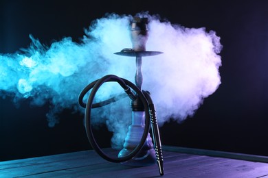 Photo of Hookah on wooden table with smoke in color lights against black background