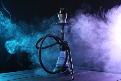 Photo of Hookah on wooden table with smoke in color lights against black background
