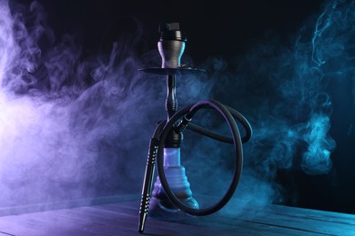 Photo of Hookah on wooden table with smoke in color lights against black background