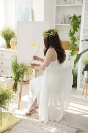 Photo of Beautiful woman with chamomile wreath drawing on canvas indoors