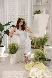 Photo of Beautiful woman with chamomile wreath near easel indoors