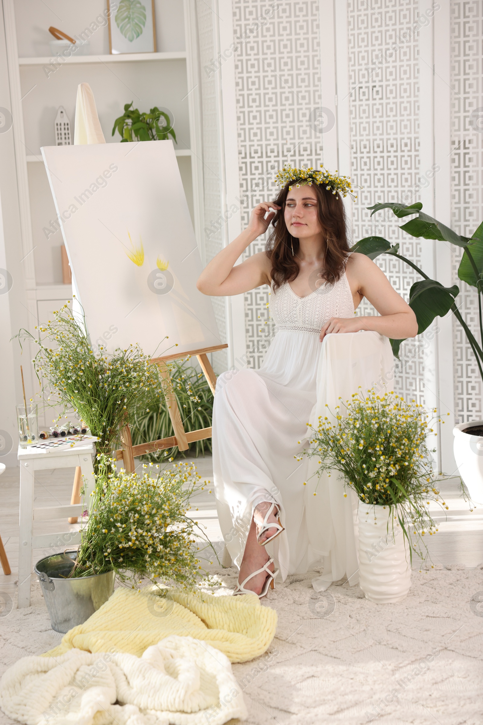 Beautiful woman with chamomile wreath near easel indoors Photo of Beautiful woman with chamomile wreath near easel indoors