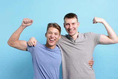 Photo of Two friends hugging and showing their biceps on light blue background