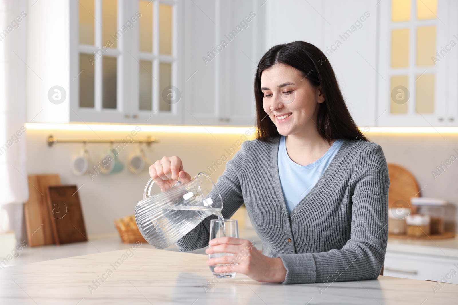 Woman pouring water into glass at white marble table in kitchen, space for text Photo of Woman pouring water into glass at white marble table in kitchen, space for text