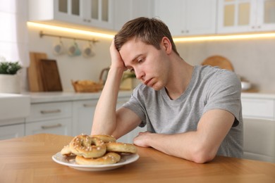 Photo of Gluten free diet. Sad woman with pretzels at wooden table in kitchen