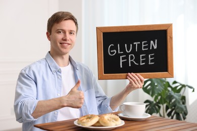 Photo of Man holding small chalkboard with text Gluten Free and showing thumbs up at wooden table indoors