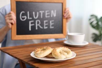 Photo of Man holding small chalkboard with text Gluten Free at wooden table indoors, focus on pretzels