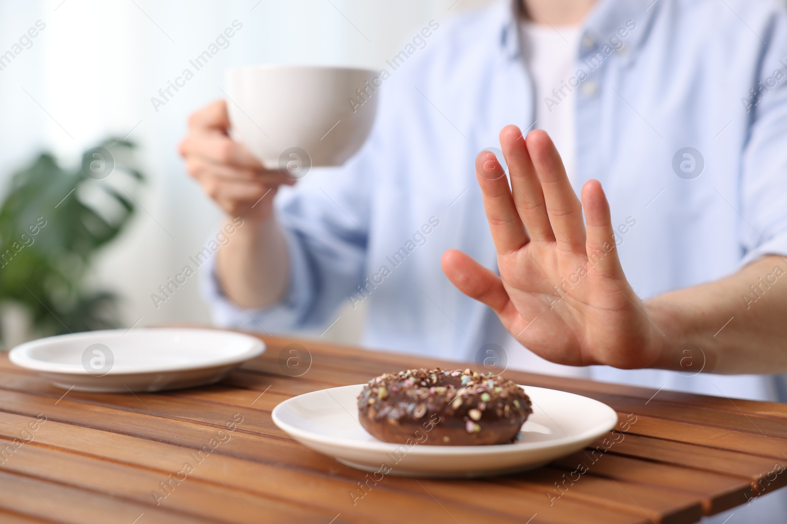 Gluten free diet. Man with cup of drink refusing from donut at wooden table indoors, closeup Photo of Gluten free diet. Man with cup of drink refusing from donut at wooden table indoors, closeup