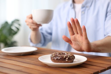 Photo of Gluten free diet. Man with cup of drink refusing from donut at wooden table indoors, closeup