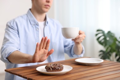 Photo of Gluten free diet. Man with cup of drink refusing from donut at wooden table indoors, closeup