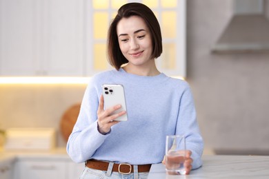 Photo of Woman with glass of water using modern smartphone at table in kitchen