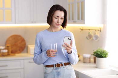 Photo of Woman with glass of water using modern smartphone in kitchen