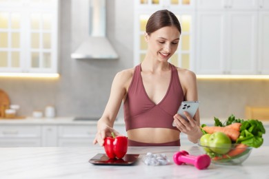 Photo of Woman with smartphone weighting bell pepper on kitchen scale at white marble table indoors