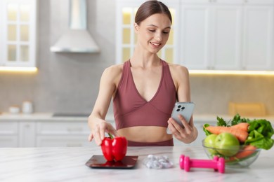 Photo of Woman with smartphone weighting bell pepper on kitchen scale at white marble table indoors